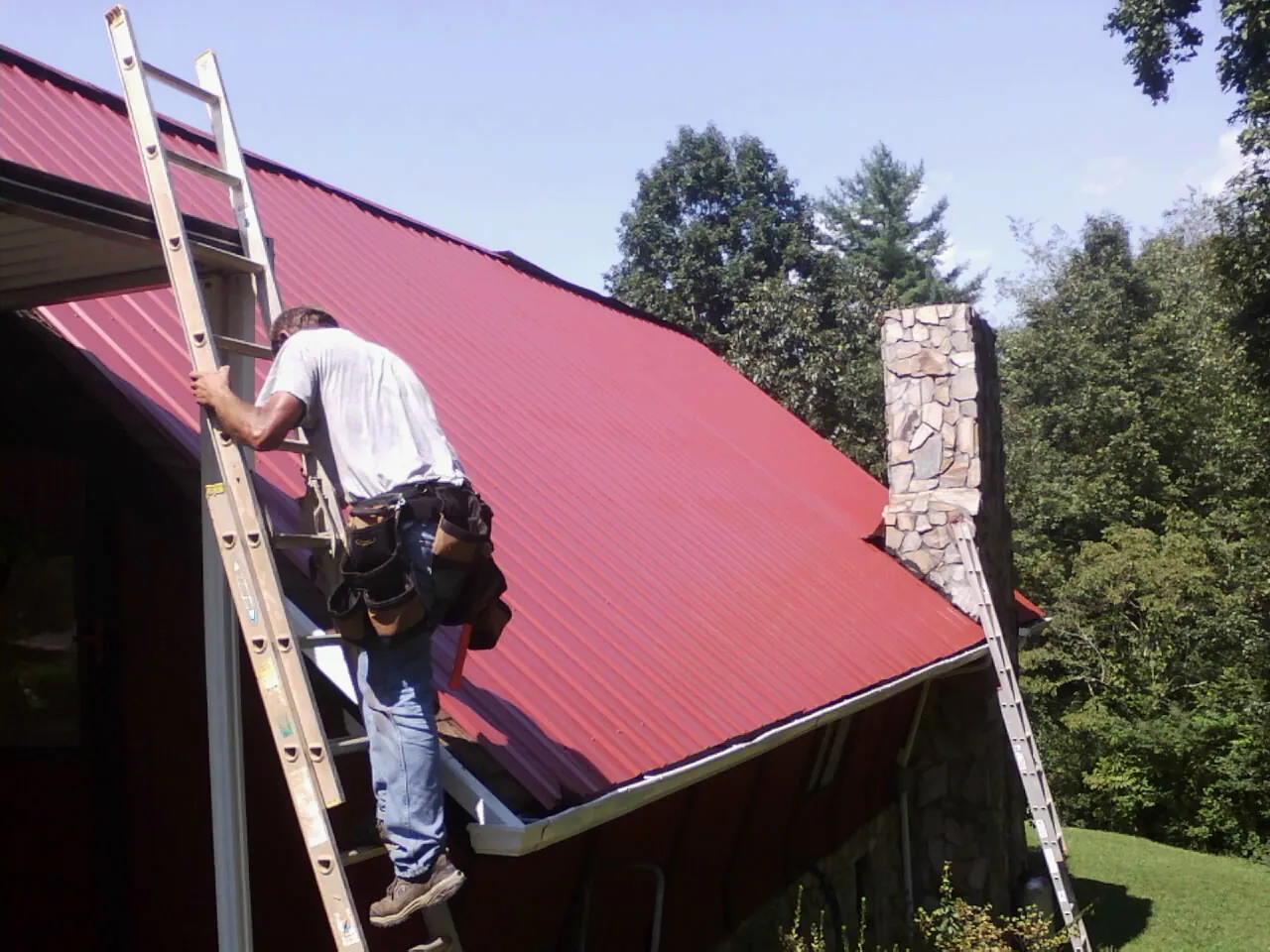 Roofer on a ladder beside a red metal roof and stone chimney — Soffit & Fascia Repair by Roofing Brothers in Accokeek